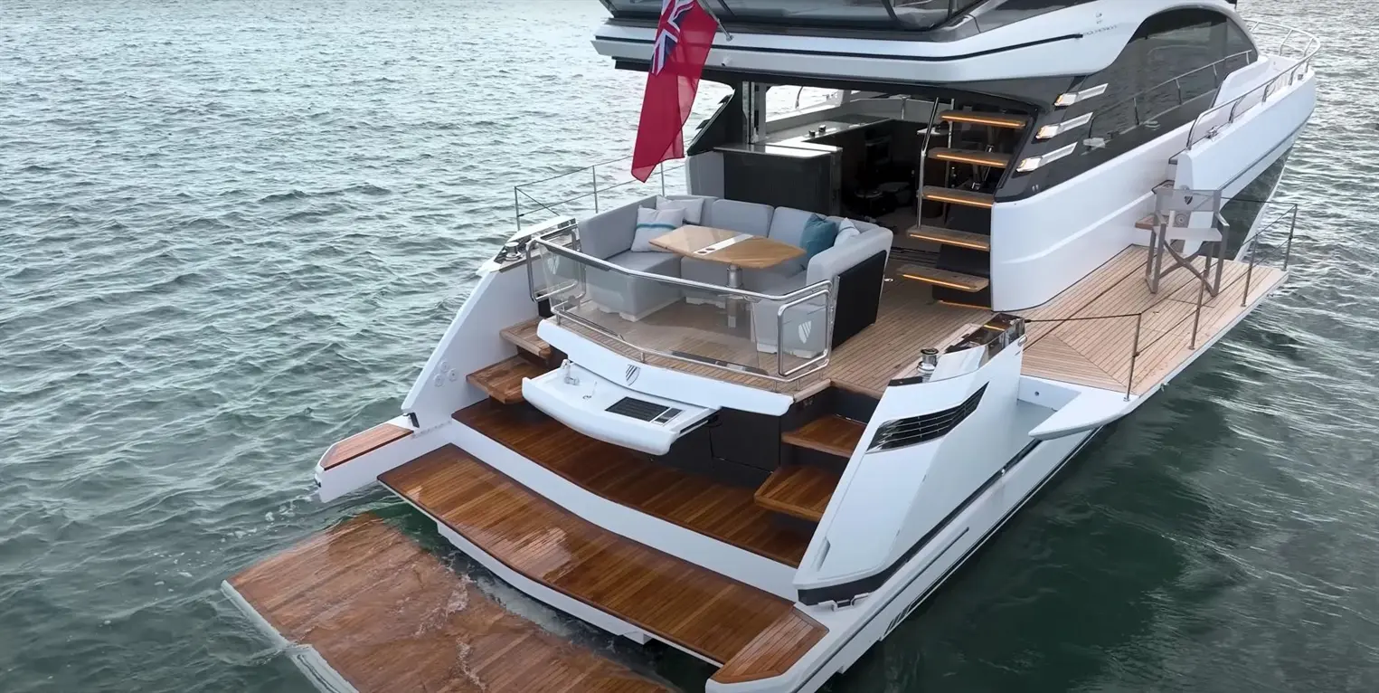 A luxury yacht with a spacious deck, large lounge area, and wooden steps on tranquil water, featuring a flag at the rear.